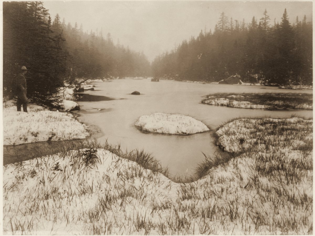 A man stands at the snowy edge of Lake Tear of the Clouds in 1888, looking out over the small pond toward the spruce on the far shore.